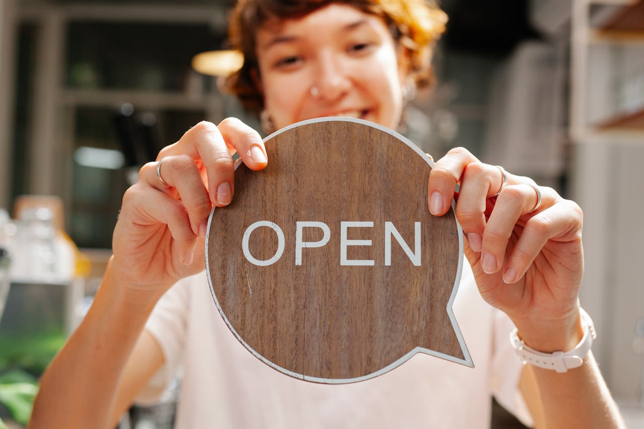 creative-03 Happy young woman worker in casual clothes standing in light cafe and showing small sign with word open